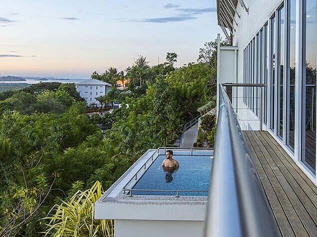 Looking down from a balcony over tropical gardens to a swimmer in a small pool, with the bay beyond