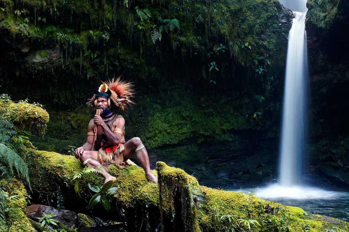 A Huli man in a feathered wig playing a pipe on a mossy log beside a rainforest waterfall