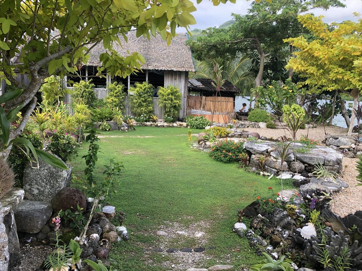 A lawn and flowering garden edged with stones and shells in front of a thatched house