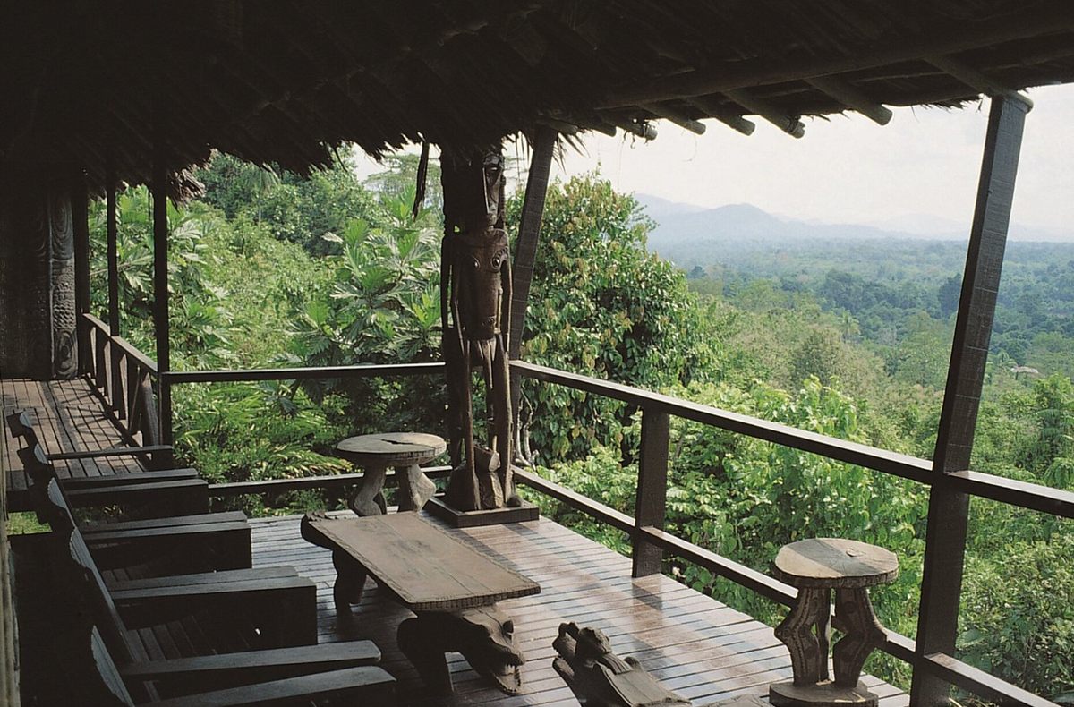 A carved figure and timber furniture on the lodge verandah, looking out over the rainforest to distant hills