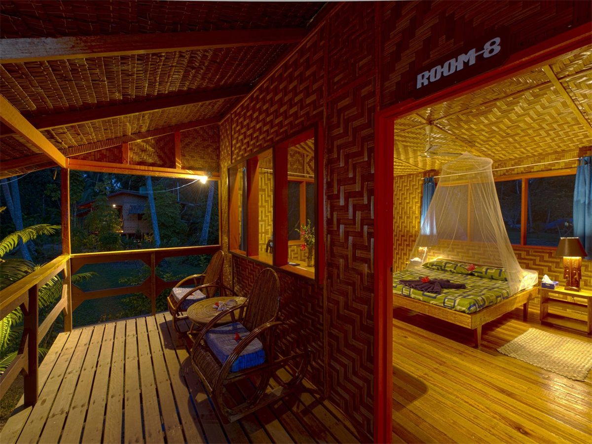 A Lissenung bungalow room with woven walls and a mosquito-netted bed, opening onto a timber verandah with chairs