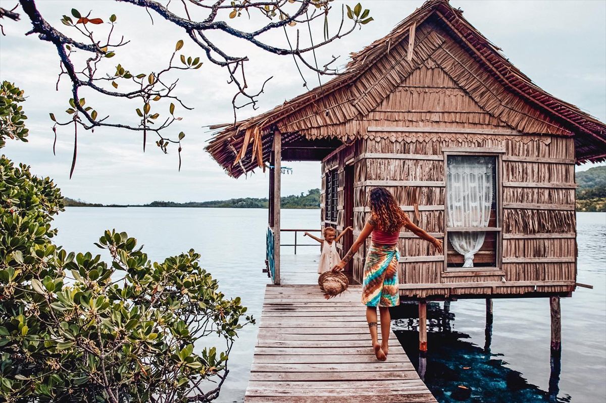 A woman and a small child on the boardwalk outside a leaf-thatched bungalow on stilts over the water