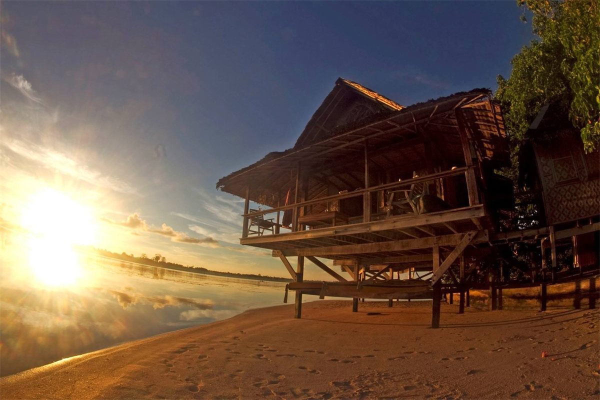 A thatched bungalow raised on stilts at the water’s edge at Nusa Island Retreat, with the sun low behind it