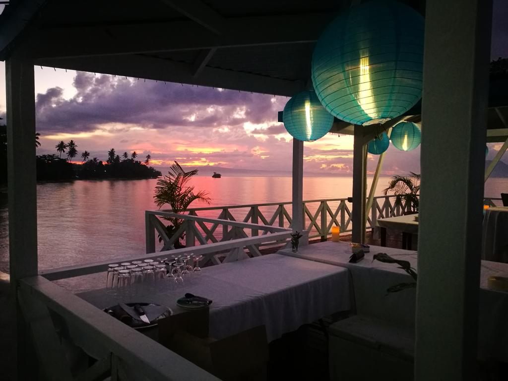 Tables set under paper lanterns on the waterfront verandah at Rapopo, with the bay at sunset beyond