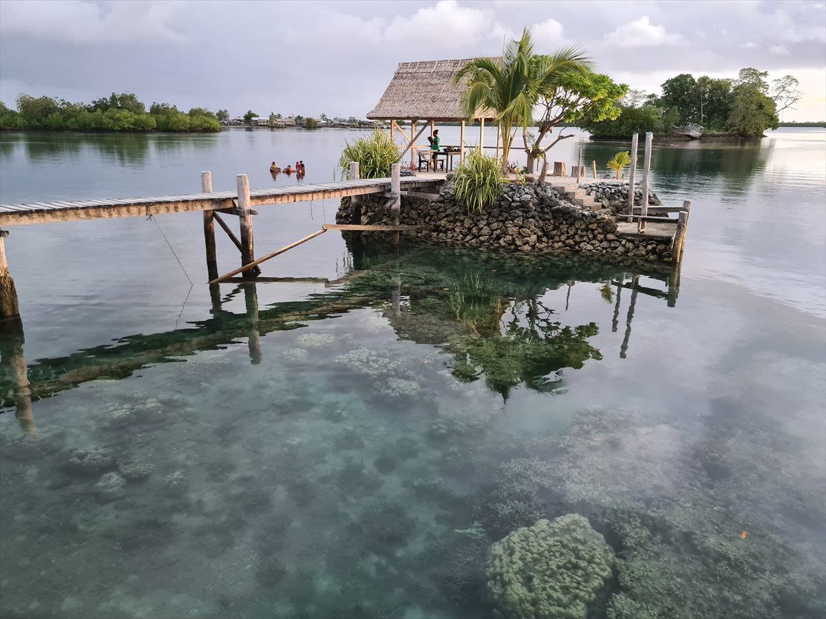 A thatched shelter on a man-made island of piled coral stone, with a jetty and coral visible through the clear water