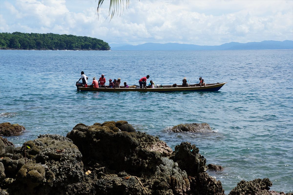 Villagers in a long motor canoe off a rocky shore, with forested islands behind