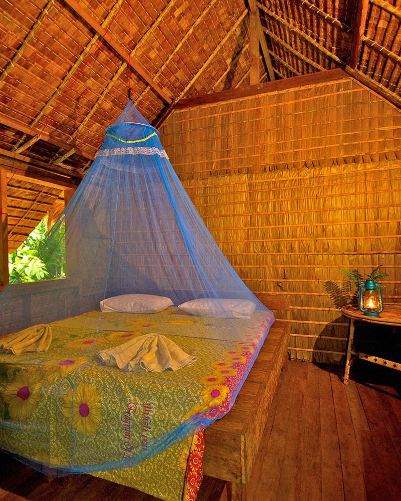 A double bed under a mosquito net inside a leaf haus with woven walls and a lantern